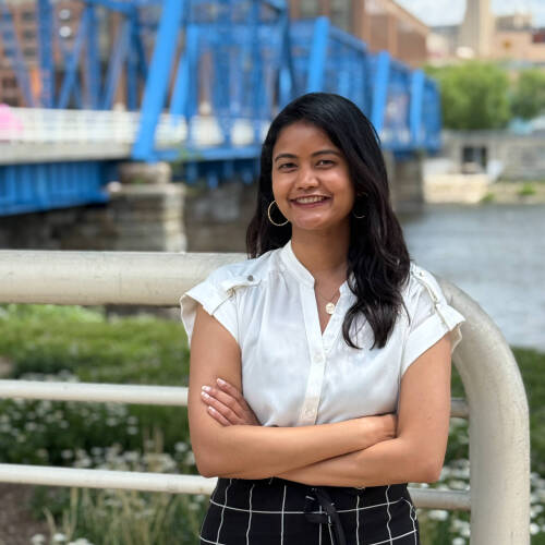 A lady with long dark hair is standing outdoors in front of a blue metal bridge, with her arms crossed and dressed in a white short-sleeve blouse and a black skirt with a white grid pattern. The background includes green grass, flowers, and city buildings